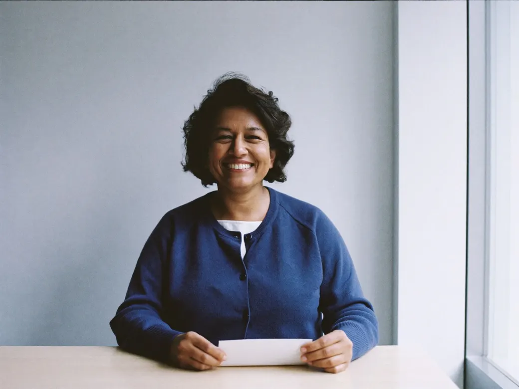 A woman in her late forties smiles holding a sheet of paper, seated by a window